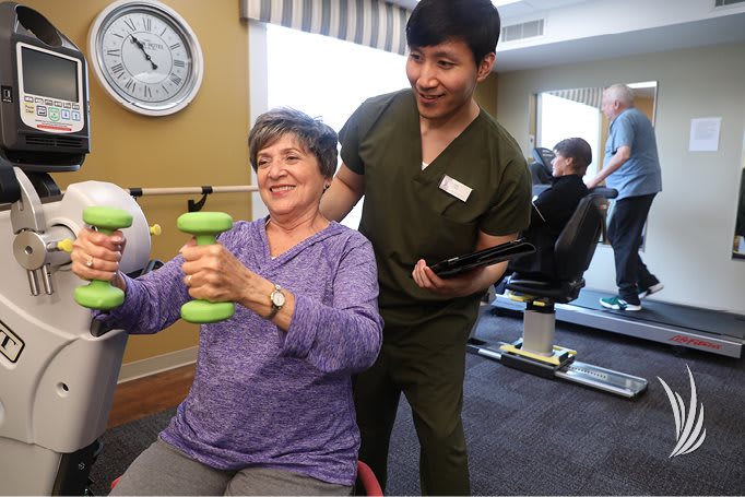 A senior woman exercising with the assistance of a caregiver in a fitness or rehabilitation setting.