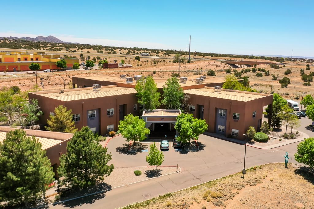 Aerial view of a building situated in a desert landscape with surrounding greenery and parking area.