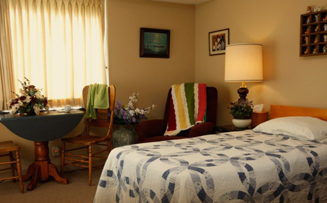 A cozy bedroom with a decorated bed, chairs by the window, and a lamp on the nightstand.