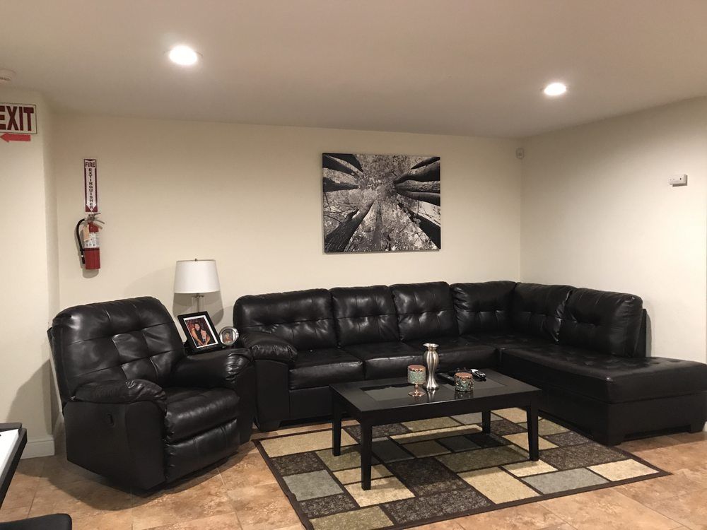 A cozy living room featuring a black leather sectional sofa and armchair, complemented by a modern coffee table and decorative items on a patterned rug. Wall art adds character to the space.