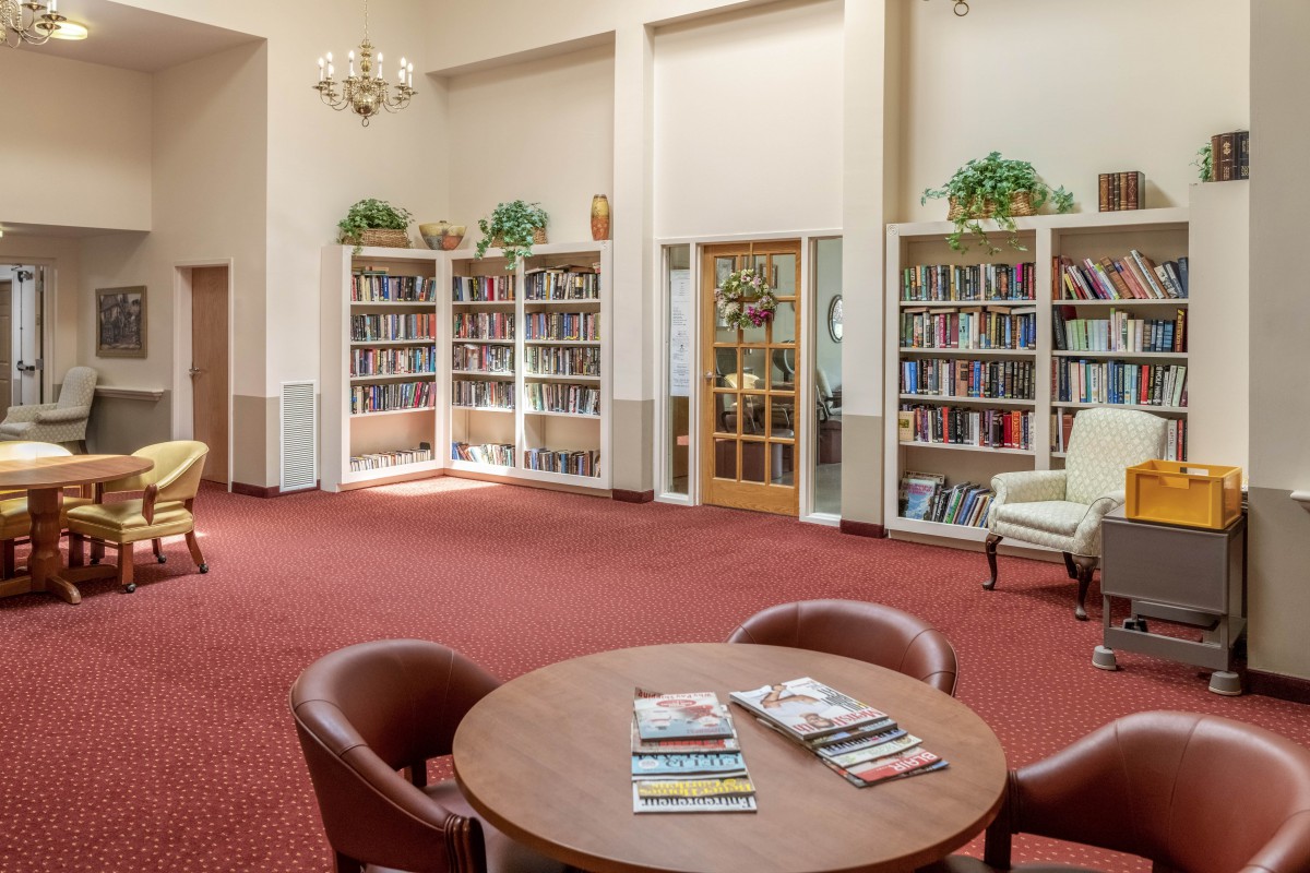 A cozy library reading room with bookshelves, armchairs, and a round table with magazines.
