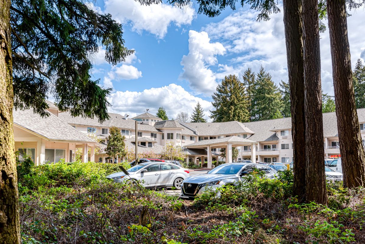 A scenic view of a building surrounded by trees with cars parked in front under a blue sky with clouds.