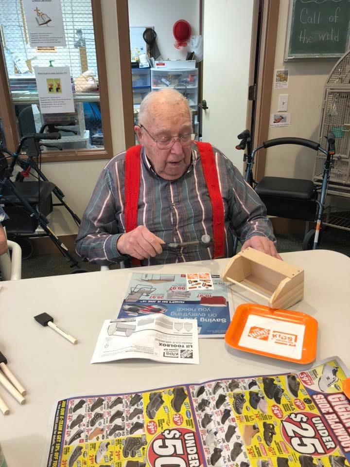 An elderly man wearing red suspenders sits at a table working on a craft project indoors.