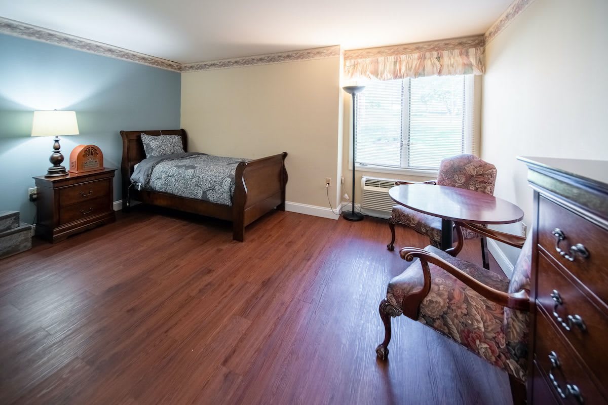 A cozy bedroom with wooden flooring, featuring a single bed, armchairs, and a table by the window.