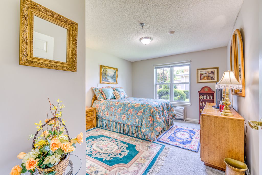 A cozy bedroom with a decorative bedspread, colorful rugs, and wall art, illuminated by natural light from a large window.