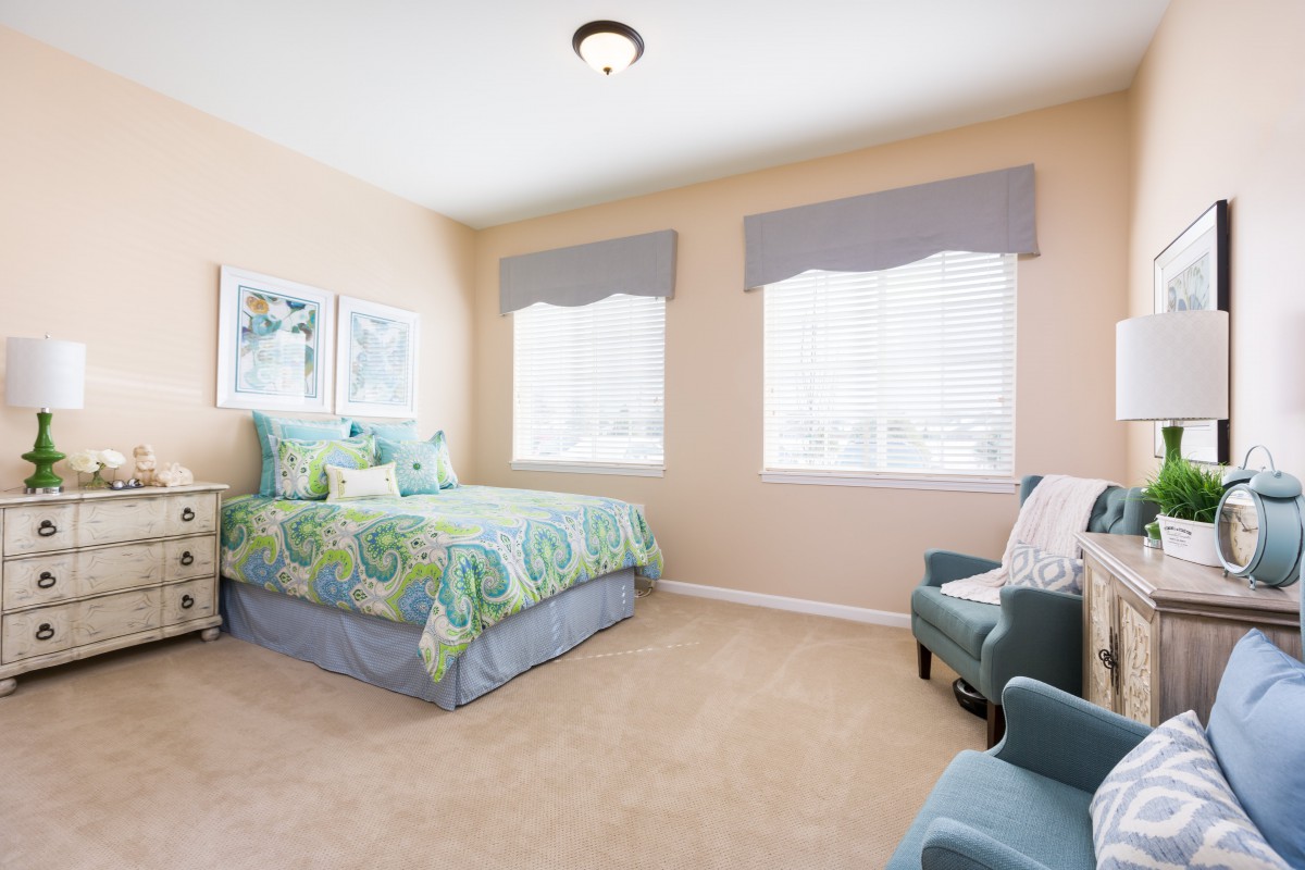 A cozy bedroom featuring a bed with colorful bedding, two windows with light curtains, and a comfortable armchair.