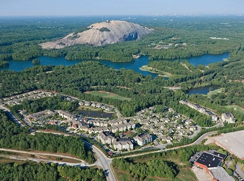 Aerial view of a residential area surrounded by lush forests and water bodies with a prominent mountain in the background.