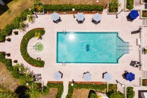 Aerial view of a well-maintained swimming pool surrounded by patio umbrellas and lush landscaping on a sunny day.