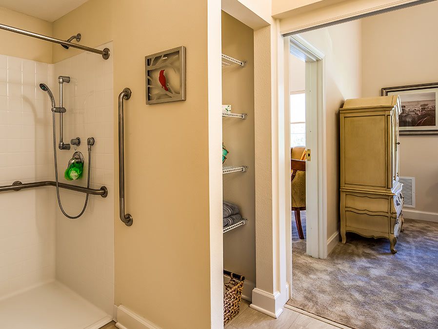 A well-lit bathroom featuring a shower with handrails and built-in storage shelves, leading to an adjacent room with furniture.
