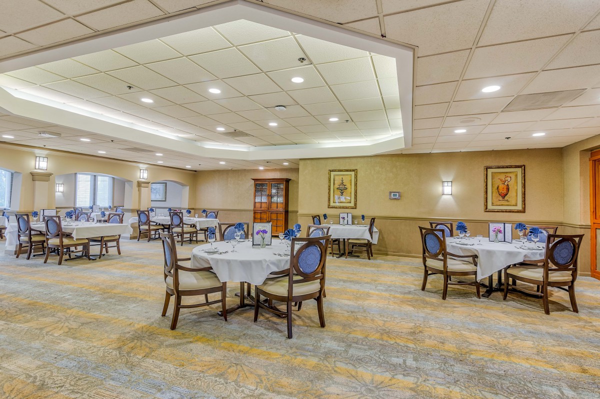 An elegant dining room with neatly arranged tables and chairs, featuring bright lighting and a carpeted floor.