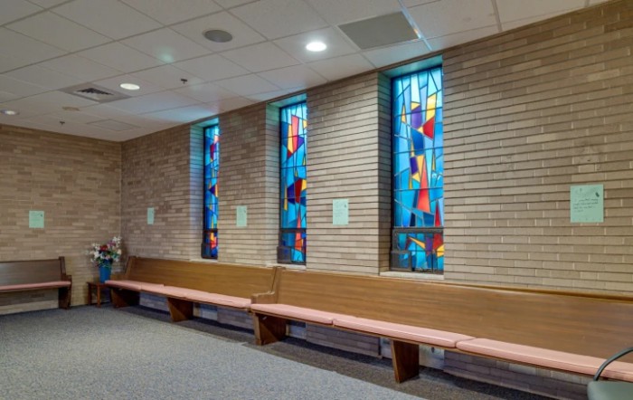 A chapel interior with stained glass windows and a wooden bench along a brick wall.