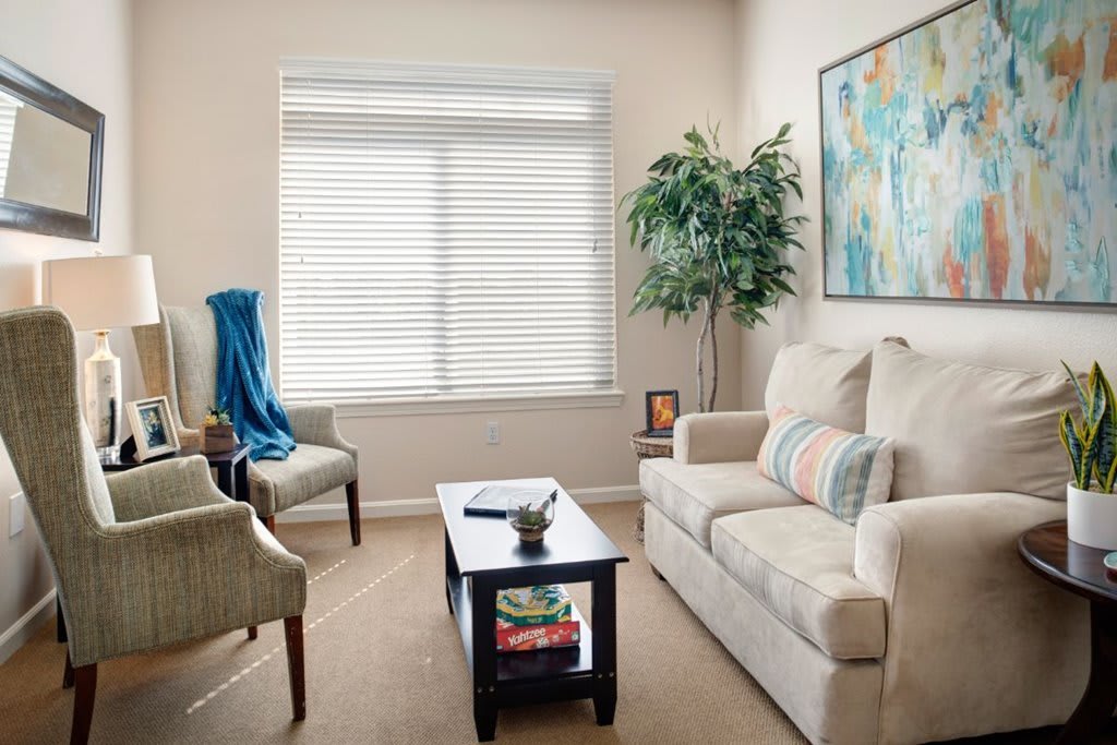 A cozy living room with a light-colored couch, armchairs, and a coffee table. The room is decorated with artwork, a potted plant, and window blinds.