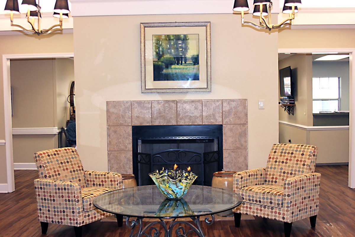 A cozy living room features a central fireplace, flanked by patterned chairs and a glass coffee table topped with decorative items.