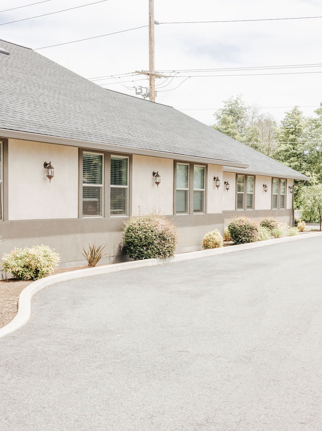 A single-story building with multiple windows, surrounded by shrubs and a paved road.