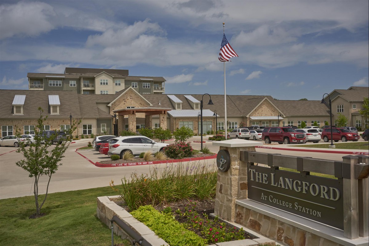 A welcoming entrance to a building complex featuring well-maintained landscaping, parked cars, and an American flag flying in the foreground.