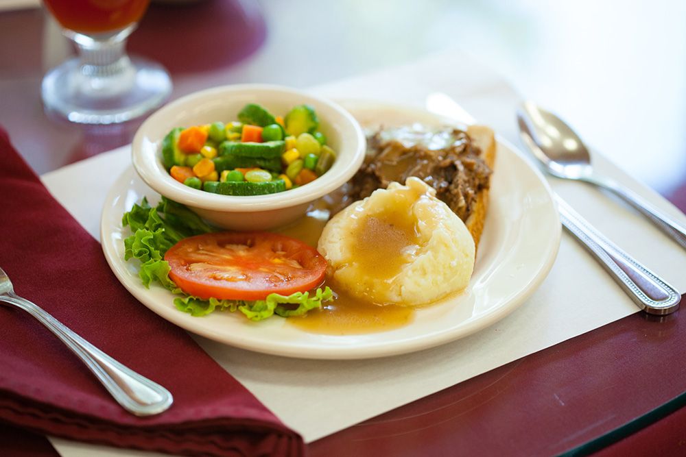 A plate featuring mashed potatoes with gravy, beef, a side of mixed vegetables, and sliced tomato.