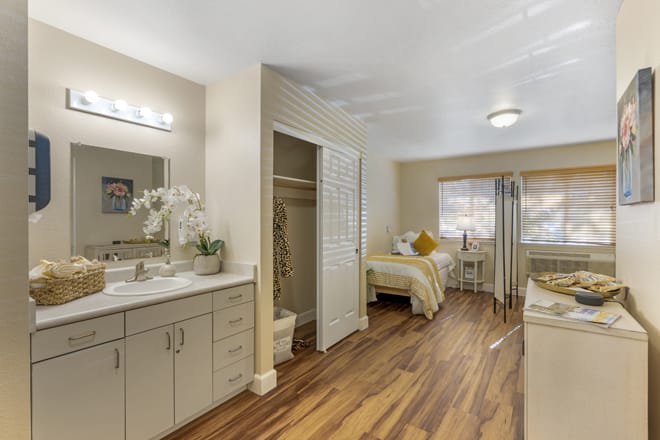 A well-decorated bedroom with wooden flooring, featuring a bed, a vanity area with a mirror, and neutral color tones.