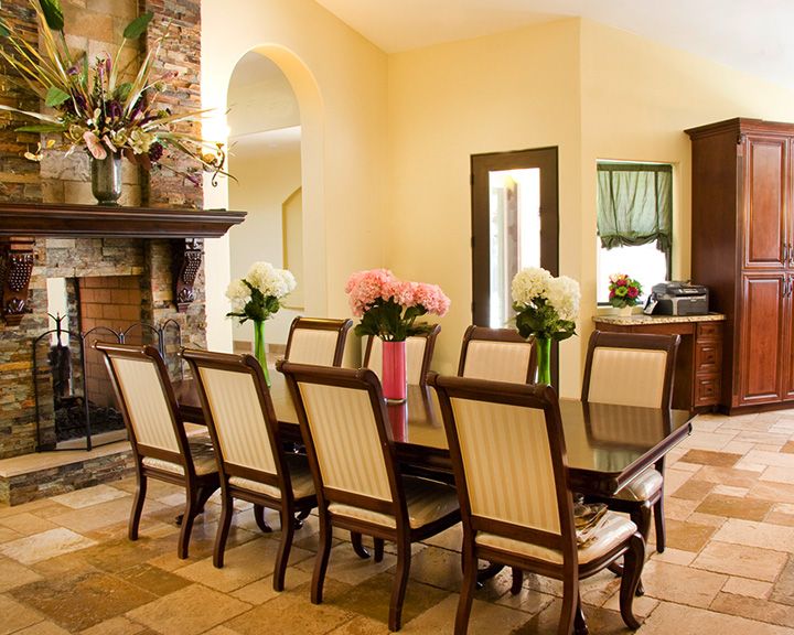 A cozy dining room with a large table surrounded by chairs, adorned with floral arrangements and featuring a stone fireplace.