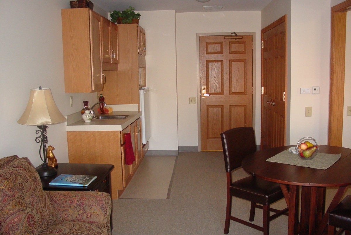 A compact apartment kitchen with a small table and chairs, showcasing wooden cabinets and a front door.