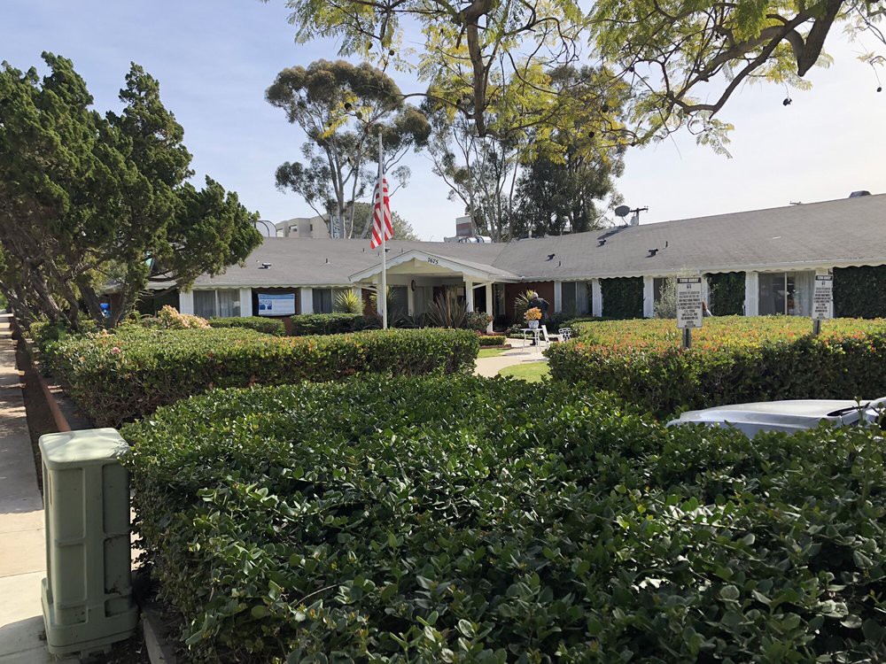 A school building surrounded by hedges and trees under a clear sky, with an American flag on a flagpole.