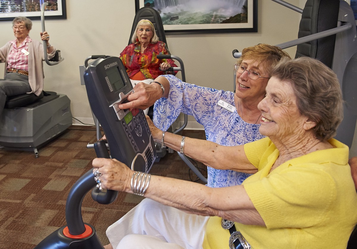 An elderly woman receives assistance while using exercise equipment in a community center.