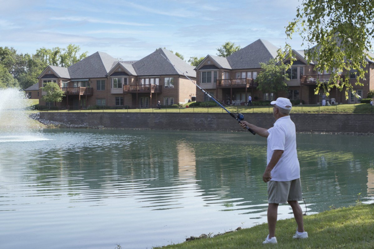 A person fishing by a lake near a residential area with several houses in the background.