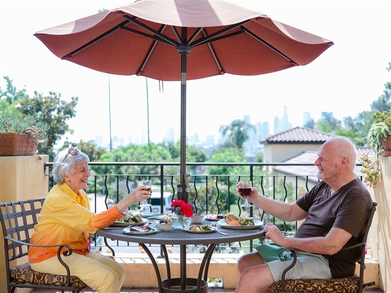 An elderly couple enjoying a meal and wine on a patio under an umbrella.