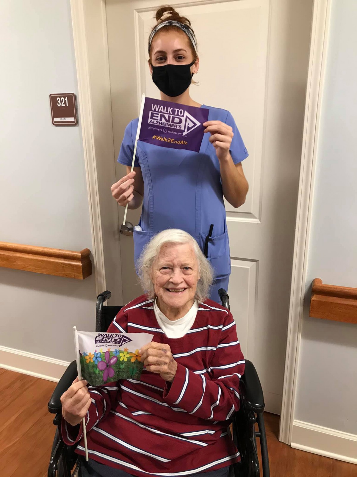 An elderly woman in a wheelchair holding a card, accompanied by a masked healthcare worker standing behind her.