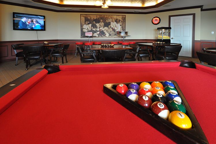 A billiards room featuring a red-felt pool table with racked balls, surrounded by tables and chairs.