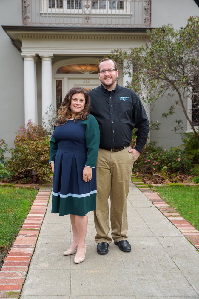 A couple posing in front of a house, dressed in formal attire.