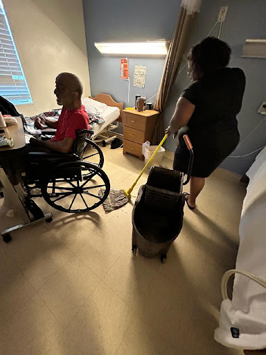 A caregiver cleans the floor in a hospital room while attending to a patient in a wheelchair.