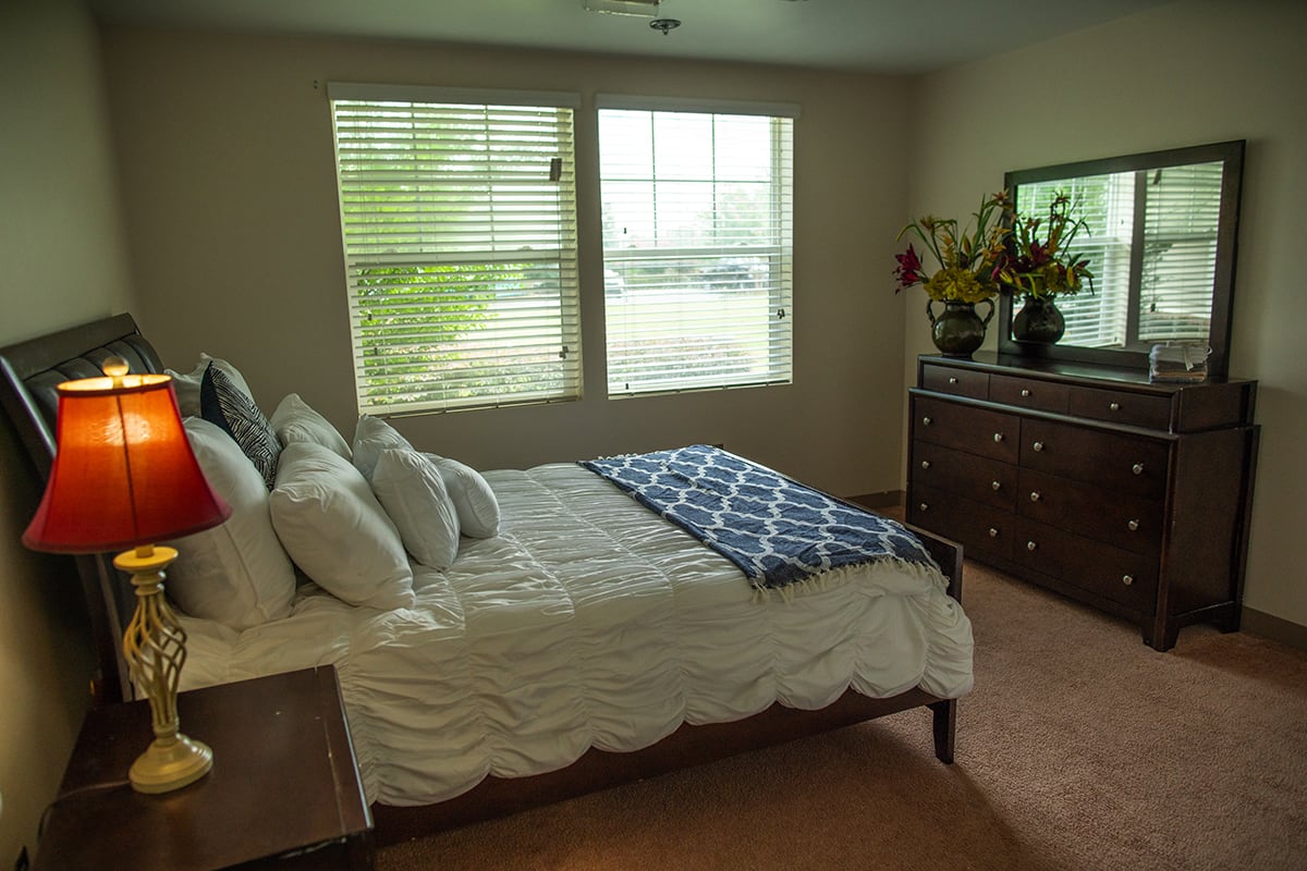 A cozy bedroom featuring a neatly made bed, a dresser with decorative items, and large windows letting in natural light.