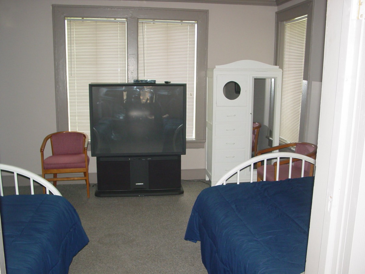A bedroom with two beds, a large television, chairs, and a mirrored wardrobe.