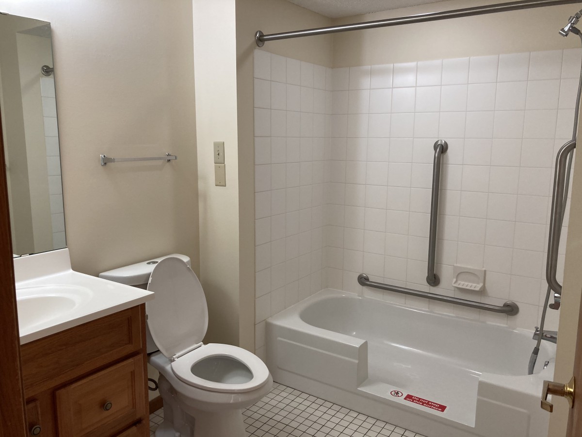 A clean, white-tiled bathroom featuring a toilet, bathtub with a shower curtain, and a wooden cabinet sink.
