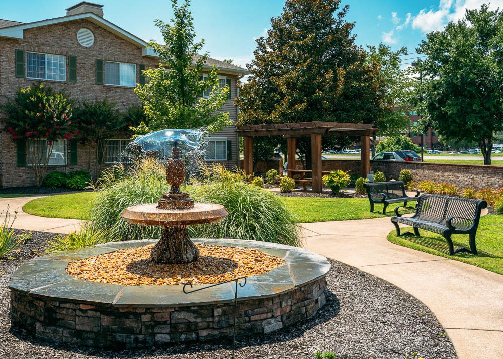 A tranquil garden setting featuring a stone fountain, benches, and lush landscaping with trees and a pathway.