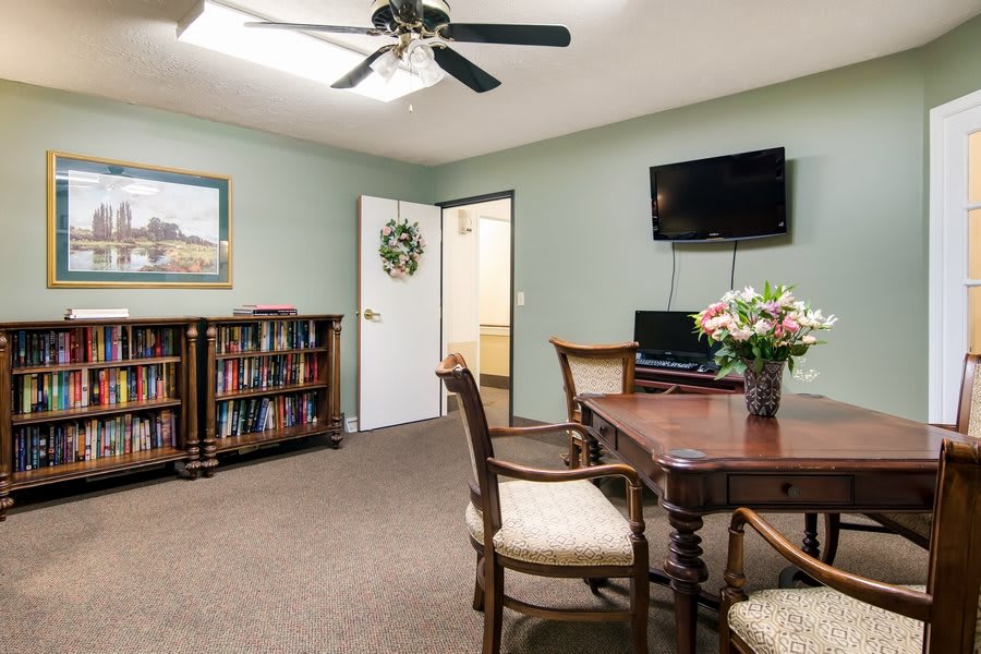 A cozy living room with a wooden table and chairs, a bookshelf, and a wall-mounted television.