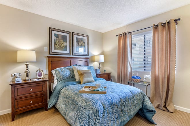 A cozy bedroom with a neatly made bed, flanked by nightstands and lamps, complemented by framed artwork above the headboard. Natural light filters through the window, enhancing the serene atmosphere.