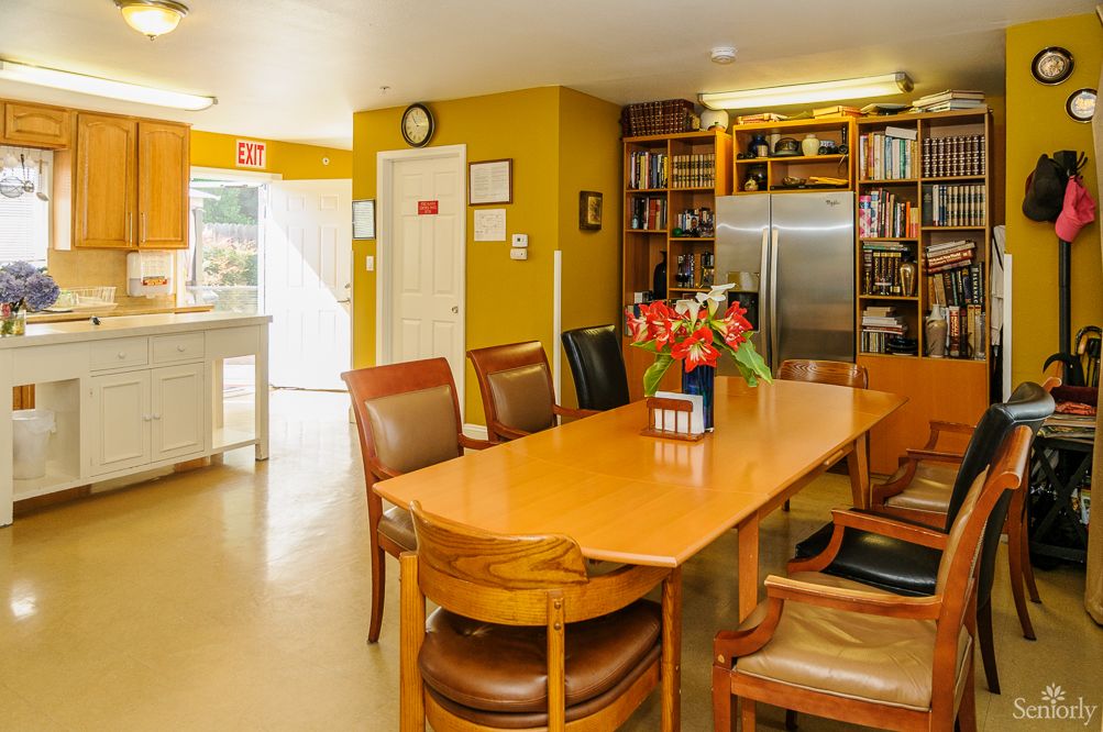 A cozy kitchen and dining area with a large wooden table, surrounded by chairs, adorned with flowers and flanked by bookshelves and a refrigerator.