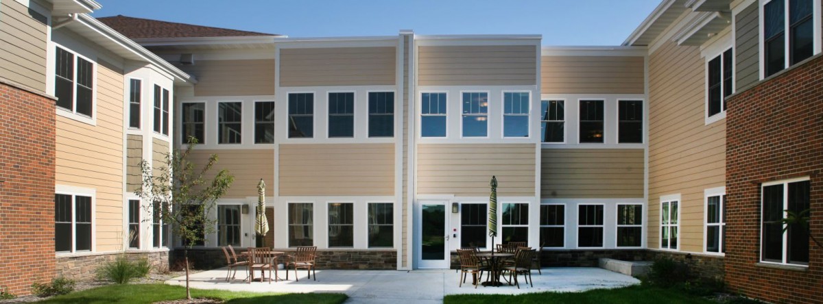 A modern two-story building with a beige exterior and large windows, featuring outdoor patio furniture.