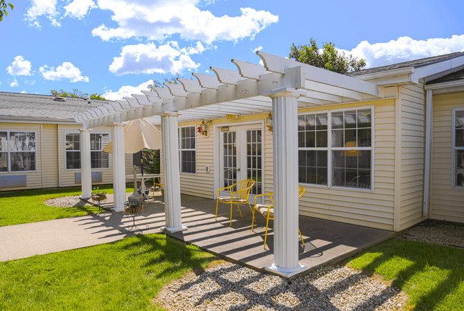 A sunny backyard featuring a white pergola attached to a house, with a patio area and green lawn.