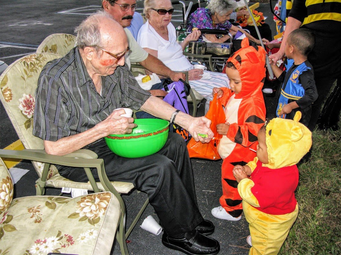 An elderly man hands out candy to children dressed in Winnie the Pooh and Tigger costumes during Halloween.