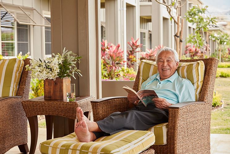 An elderly man is comfortably reading a book while relaxing on a patio chair.
