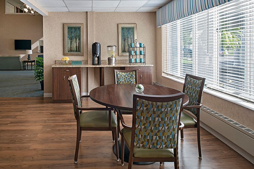 A cozy dining area with a round table surrounded by four patterned chairs, featuring large windows with blinds and decorative wall art.