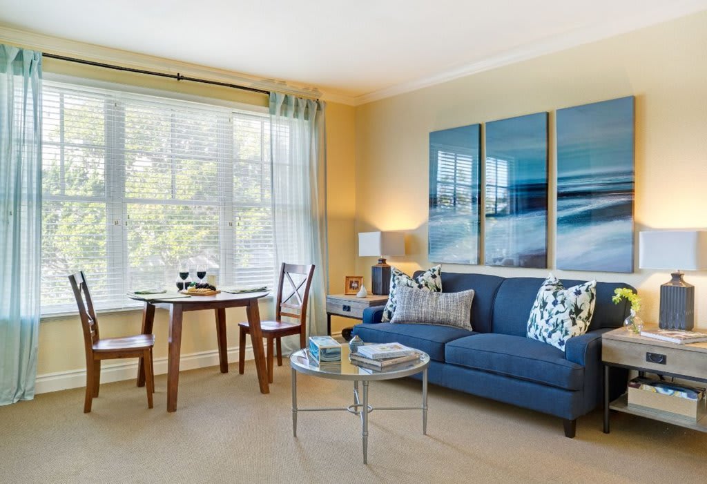 A bright and cozy living room with a blue sofa, modern artwork, and a small dining area by the window.