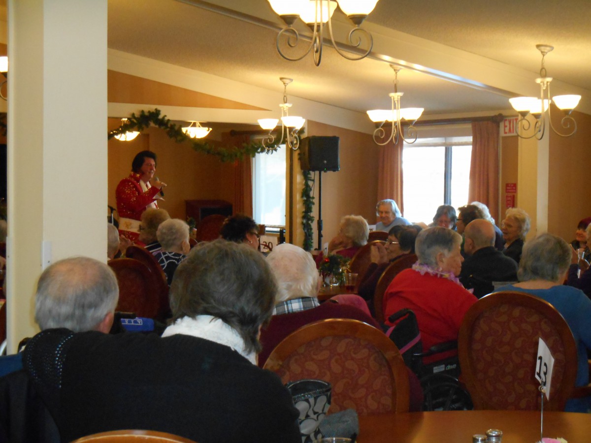A community gathering with elderly individuals listening to a speaker in an indoor venue.