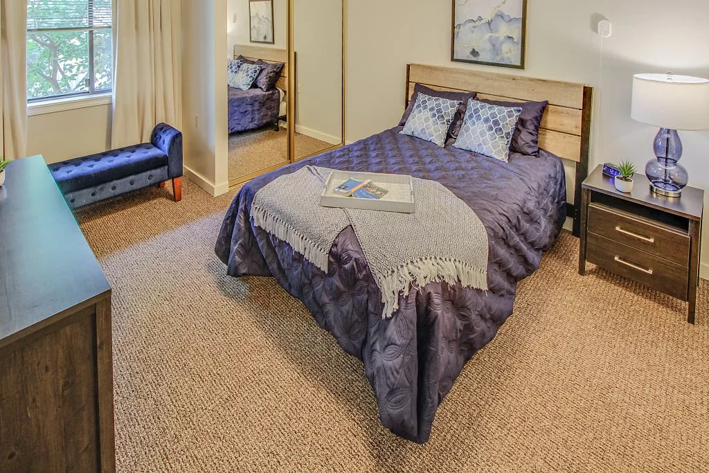 A cozy bedroom featuring a neatly made bed with decorative pillows and a comforter, a carpeted floor, and a chair in the corner.