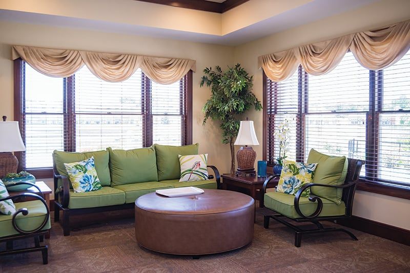 A cozy living room with green sofas, a brown round coffee table, and natural light streaming through large windows.