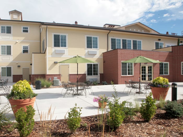 A modern building with a spacious outdoor patio featuring tables, chairs, and green umbrellas, surrounded by plants.