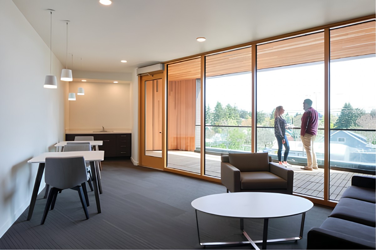 A modern office space with large glass walls, minimalist furniture, and people conversing on a balcony.