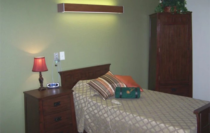 A cozy bedroom featuring a single bed with striped pillows and a quilted bedspread, accompanied by a nightstand with a lamp and an armoire against the green-painted wall.